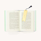The Simplified Engraved Bookmark - Magic In The Ordinary features a floral design, a yellow motif, and a navy blue tassel, shown marking the pages of an open book on a white surface.