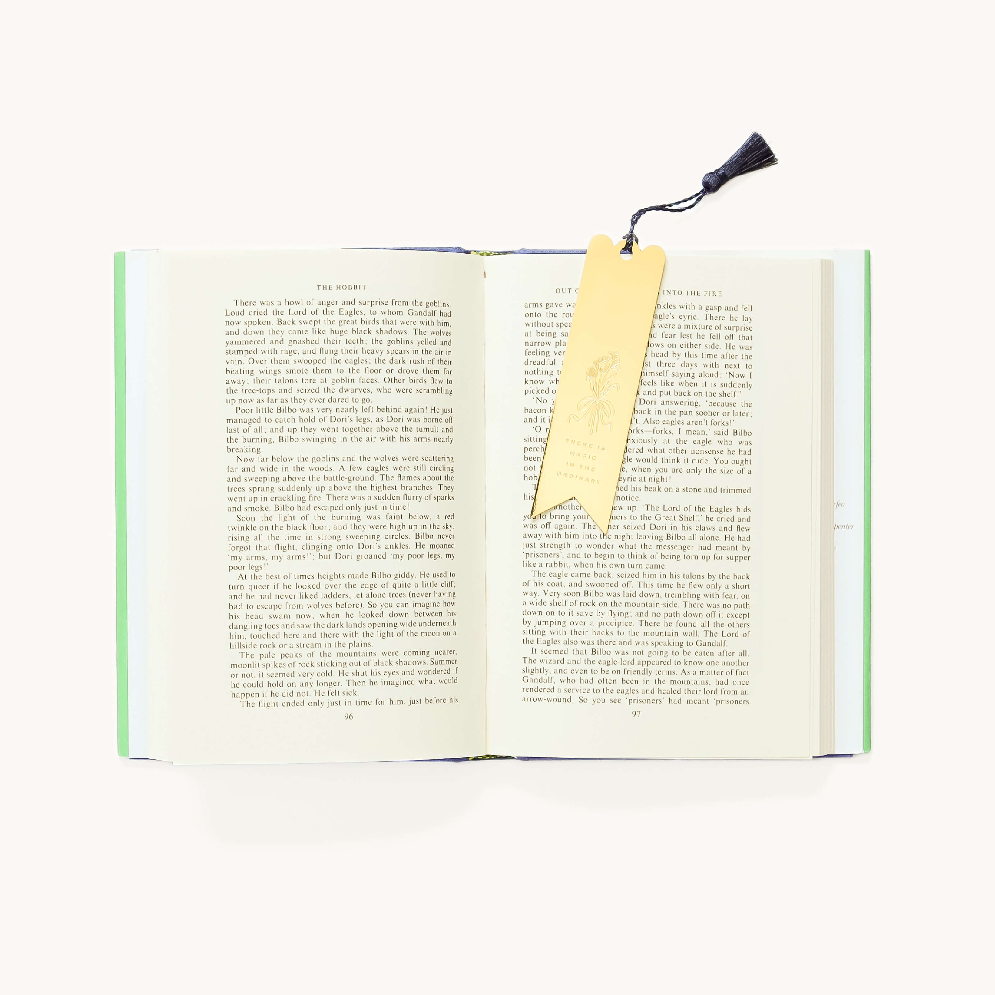 The Simplified Engraved Bookmark - Magic In The Ordinary features a floral design, a yellow motif, and a navy blue tassel, shown marking the pages of an open book on a white surface.