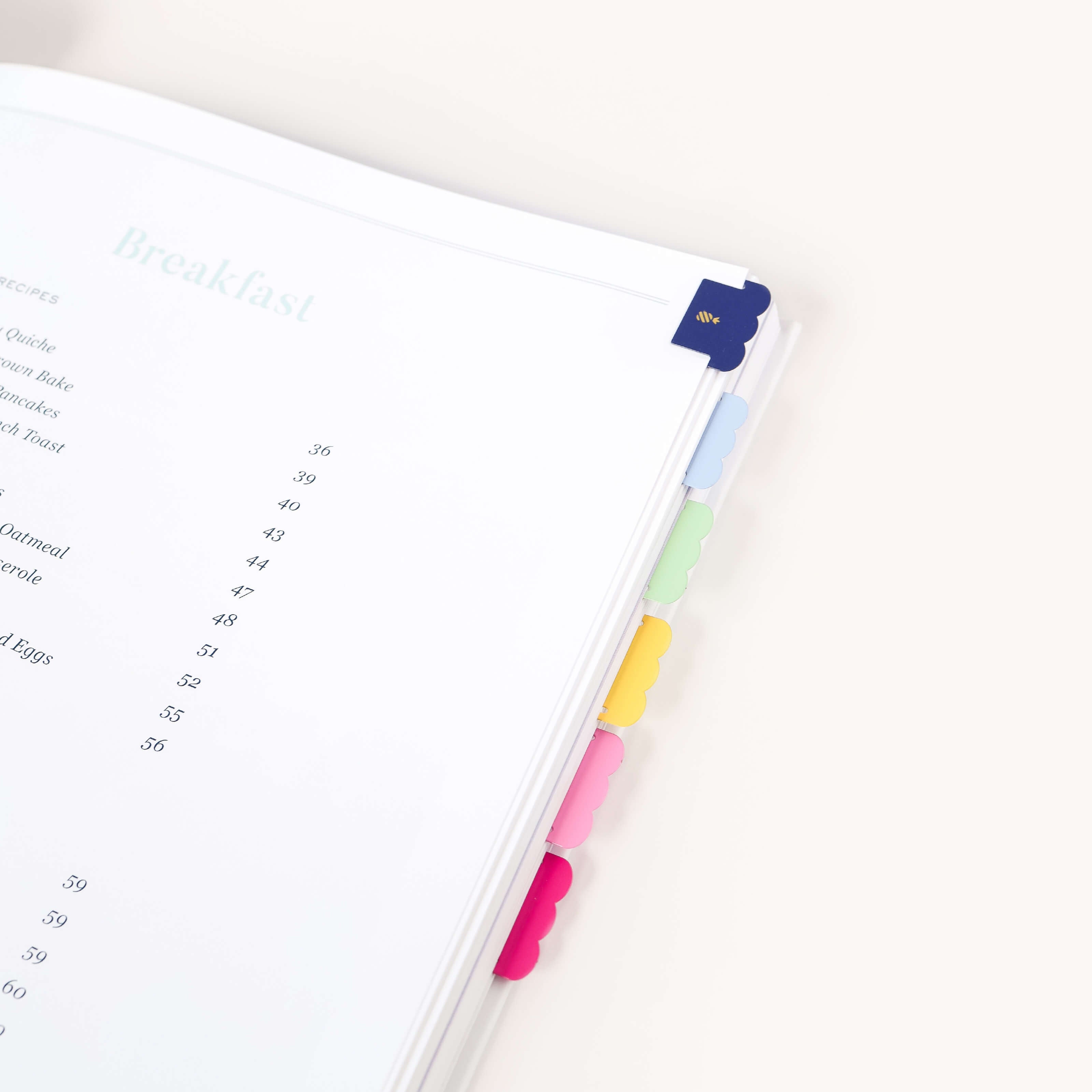 An open book with colorful planner accessories and the Simplified Index Tab Set by Simplified on the edge, opened to a breakfast section listing meal names and page numbers, set against a plain white background.
