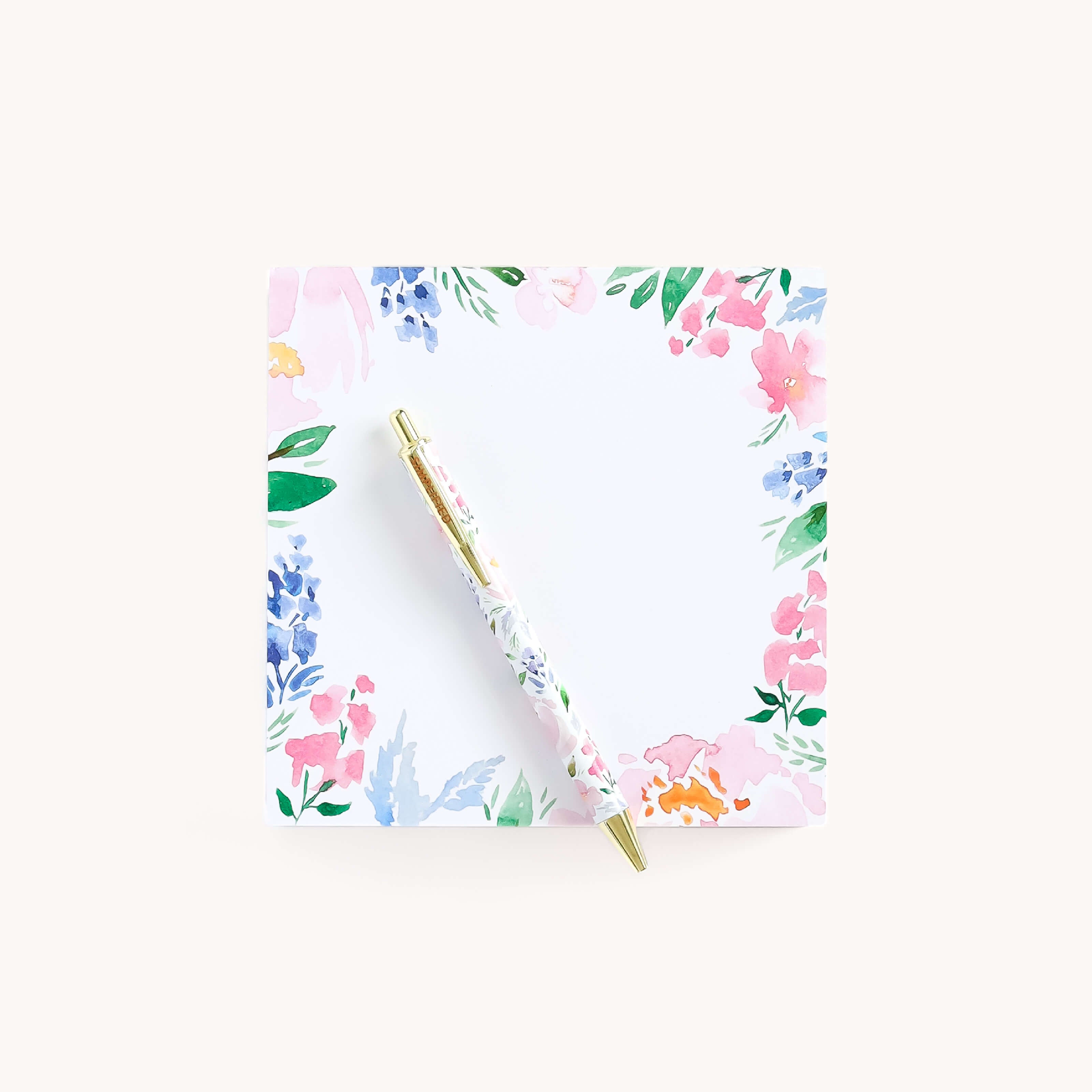 The Square Notepad – Cottage Floral by Simplified sits on a white surface, adorned with pink, blue, and green floral designs. A matching pen crosses it diagonally, creating a vibrant border and making it an ideal gift for loved ones.