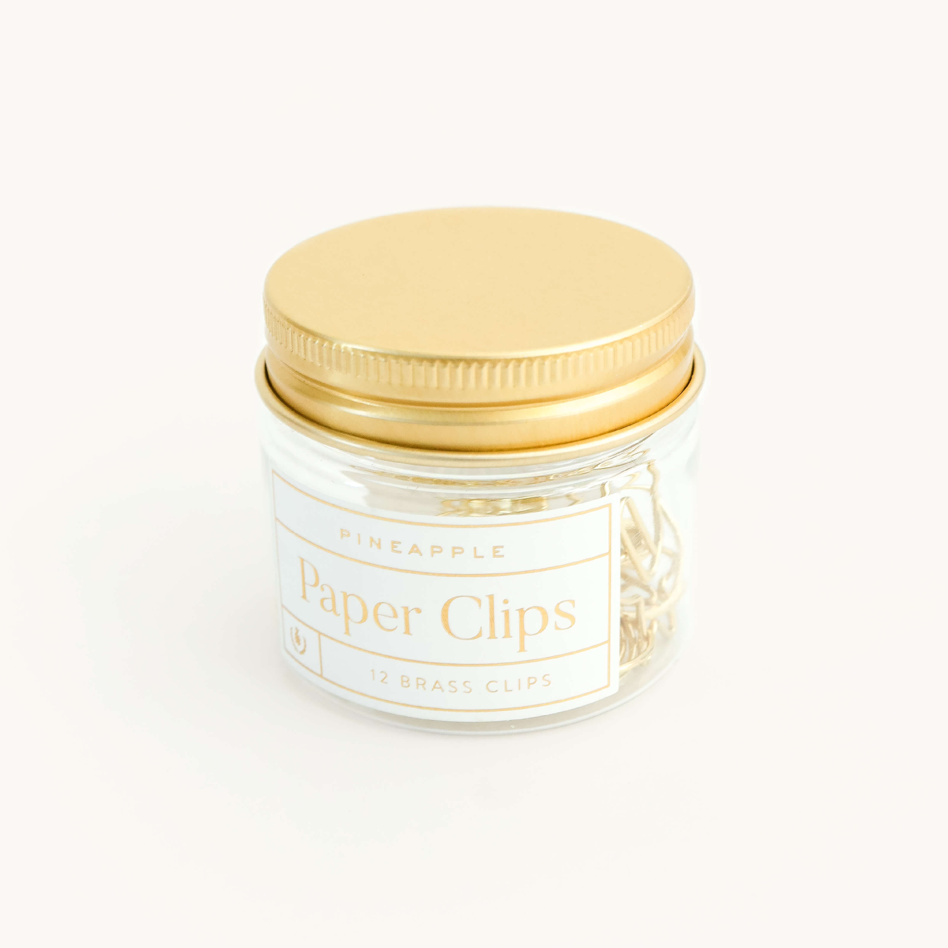 A clear glass jar with a gold lid from Simplified holds 12 brass pineapple-shaped paper clips. The label says “Paper Clips - Pineapples, 12 Brass Clips.”.