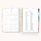 The Simplified 2026 Spiral Daily Simplified Planner - Cottage Floral features gold spiral binding and colorful tabbed sections, with a 2026 calendar on the left page and a list of 2026 holidays on the right.