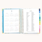 The Simplified 2026 Spiral Daily Simplified Planner – Faraway Land features a hand-painted cover, 2025 yearly calendar on the left, 2025 holidays on the right, and colorful tab dividers along the edge.