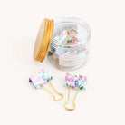A clear jar with a gold lid from Simplified contains stylish binder clips, named Cottage Floral. Two clips are outside the jar, showcasing durable gold metal handles and a colorful pink and purple floral design.