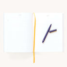 The Simplified Linen Journal - Back and Forth, with lined pages and a yellow ribbon bookmark, lies open on a plain white background. A black pen with its cap off rests on the right page, ready for your thoughts.