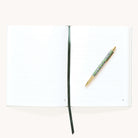 An open Simplified Linen Journal - Block Floral with a dark ribbon bookmark and a floral-patterned gold pen on the right page, set against a light background.