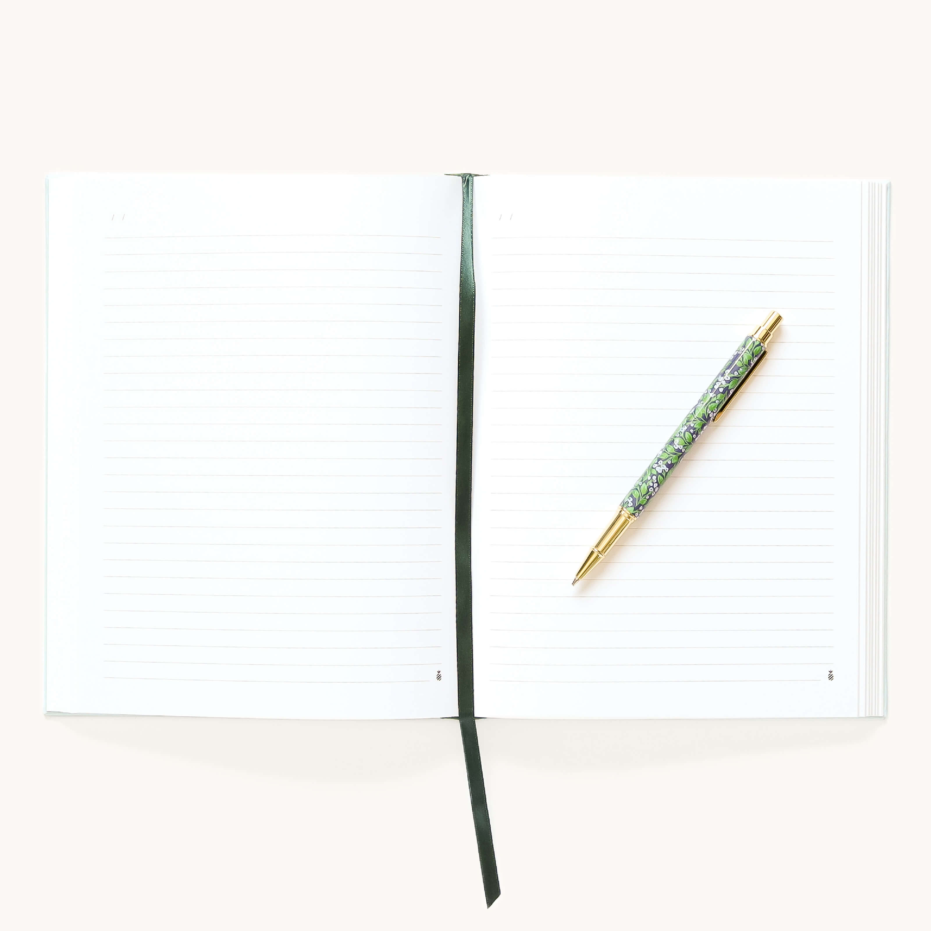 An open Simplified Linen Journal - Block Floral with a dark ribbon bookmark and a floral-patterned gold pen on the right page, set against a light background.