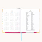 The 2026 Bookbound Weekly Simplified Planner - Happy Stripe by Simplified features lay-flat binding, a yearly calendar on the left, 2025 holidays on the right, and is accented with a blue ribbon bookmark at the bottom center.