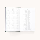 The Simplified 2026 Bookbound Daily Dapperdesk Planner - Evergreen shows a year calendar on the left and holidays with dates on the right, featuring a light background and a chic leatherette cover.