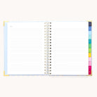 An open 2026 Spiral Weekly Simplified Planner - Serene Stripe by Simplified features lined pages, gold corner protectors, and colorful tab dividers on the right, displayed against a light background.