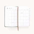 The 2026 Bookbound Daily Dapperdesk Planner - Evergreen by Simplified lies open, showing January’s monthly calendar on its leatherette cover pages with a brown ribbon bookmark and visible side tabs for other months. Most pages appear blank.