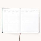 The Simplified 2026 Bookbound Weekly Dapperdesk Planner in Evergreen lies open, showing January 5–11 with daily columns, lined note sections, and a brown ribbon bookmark resting on its elegant leatherette cover.