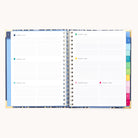 The 2026 Spiral Weekly Simplified Planner - Poppy Posies by Simplified is open to a weekly layout with lined sections for each day, colorful tab dividers on the right, and elegant gold corner protectors.
