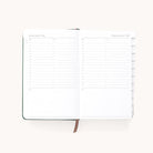 The Simplified 2026 Bookbound Daily Dapperdesk Planner in Evergreen features a leatherette cover, two-page daily spreads with hourly slots (7am–7pm), note sections, tab dividers, and a brown ribbon bookmark. Perfect for organized planning all year.