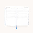 The Simplified 2026 Bookbound Daily Dapperdesk Planner - Carolina Dot features an open leatherette cover showing dated pages for Jan. 2-3, 2026, hourly schedules from 7 a.m.–8 p.m., note sections, and a blue ribbon bookmark.