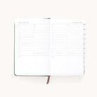 The Simplified 2026 Bookbound Daily Dapperdesk Planner in Evergreen features a leatherette cover, white pages with minimalist black text, daily schedules from 6 a.m. to 8 p.m., note sections, and tabbed monthly dividers for easy organization.