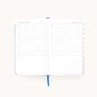 The Simplified 2026 Bookbound Daily Dapperdesk Planner in Carolina Dot has an open leatherette cover, a two-page weekly layout with hourly time slots, columns for weekend days, and a blue ribbon bookmark.