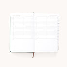 The Simplified 2026 Bookbound Daily Dapperdesk Planner in Evergreen shows two pages with hourly schedules for Saturday and Sunday. It features a leatherette cover, brown ribbon bookmark, and tabbed dividers on the right.