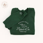 A hunter green Embroidered Crewneck - North Pole Paper Co by Simplified® features a white postage stamp graphic and "Making a list and checking it twice." A round Limited Edition tag appears in the top left corner.
