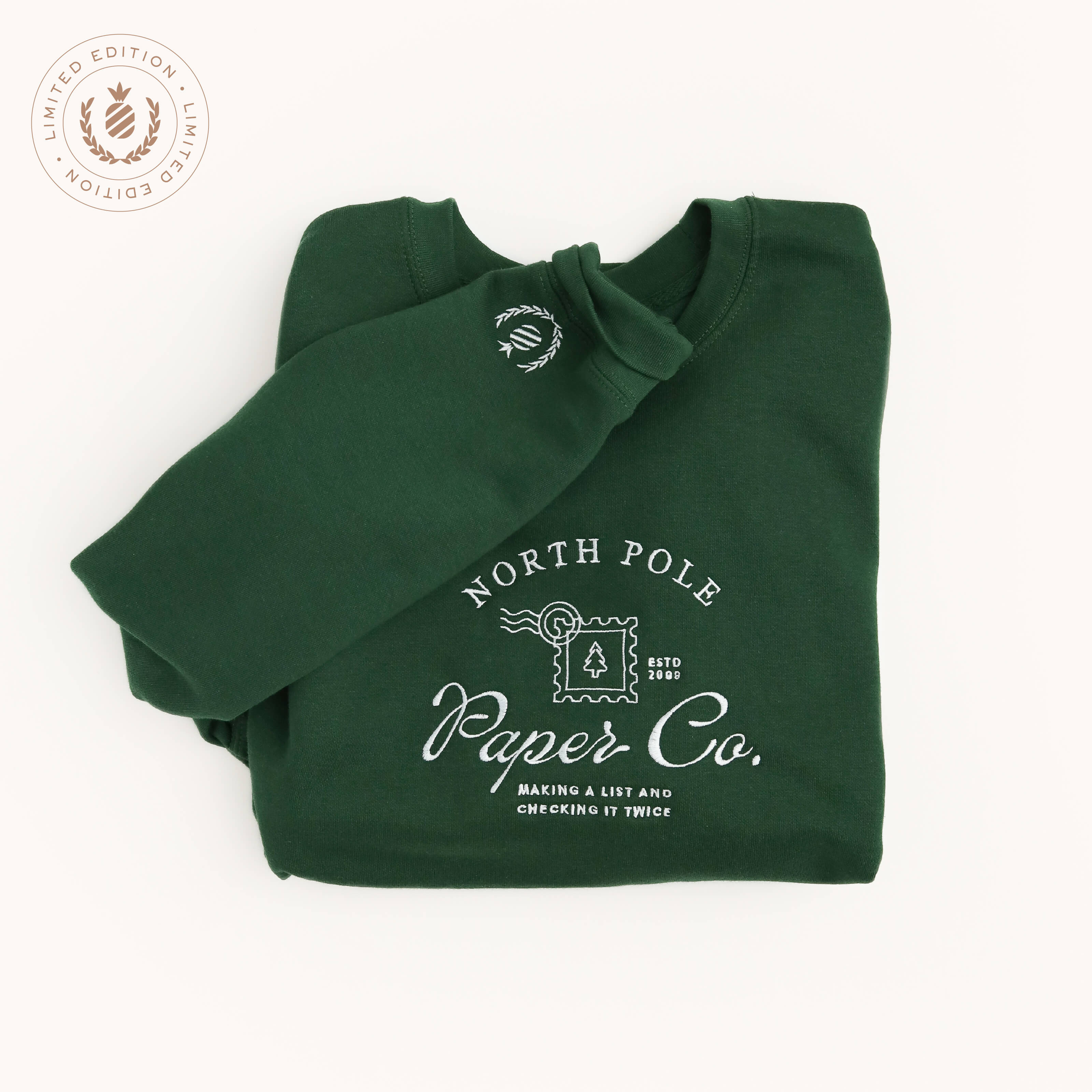 A hunter green Embroidered Crewneck - North Pole Paper Co by Simplified® features a white postage stamp graphic and "Making a list and checking it twice." A round Limited Edition tag appears in the top left corner.