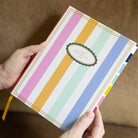 Someone holds the Simplified Linen Journal - Back and Forth, featuring pastel pink, orange, mint, blue, and white vertical stripes on its cover with colored tabs visible along the side.