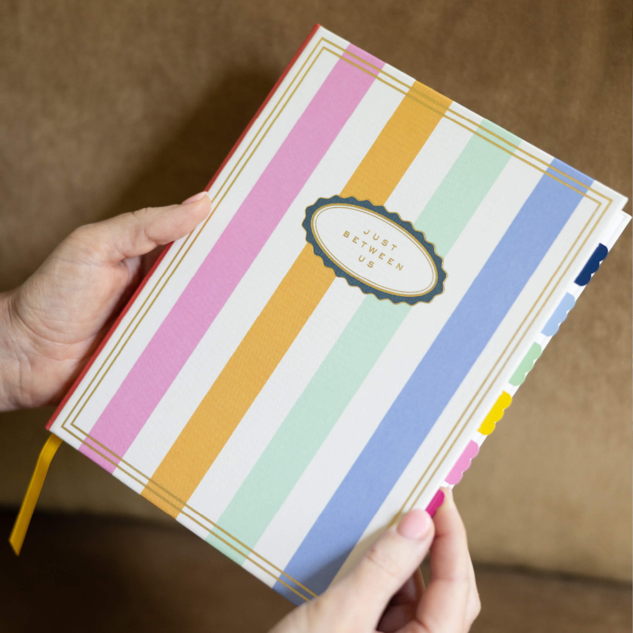 Someone holds the Simplified Linen Journal - Back and Forth, featuring pastel pink, orange, mint, blue, and white vertical stripes on its cover with colored tabs visible along the side.