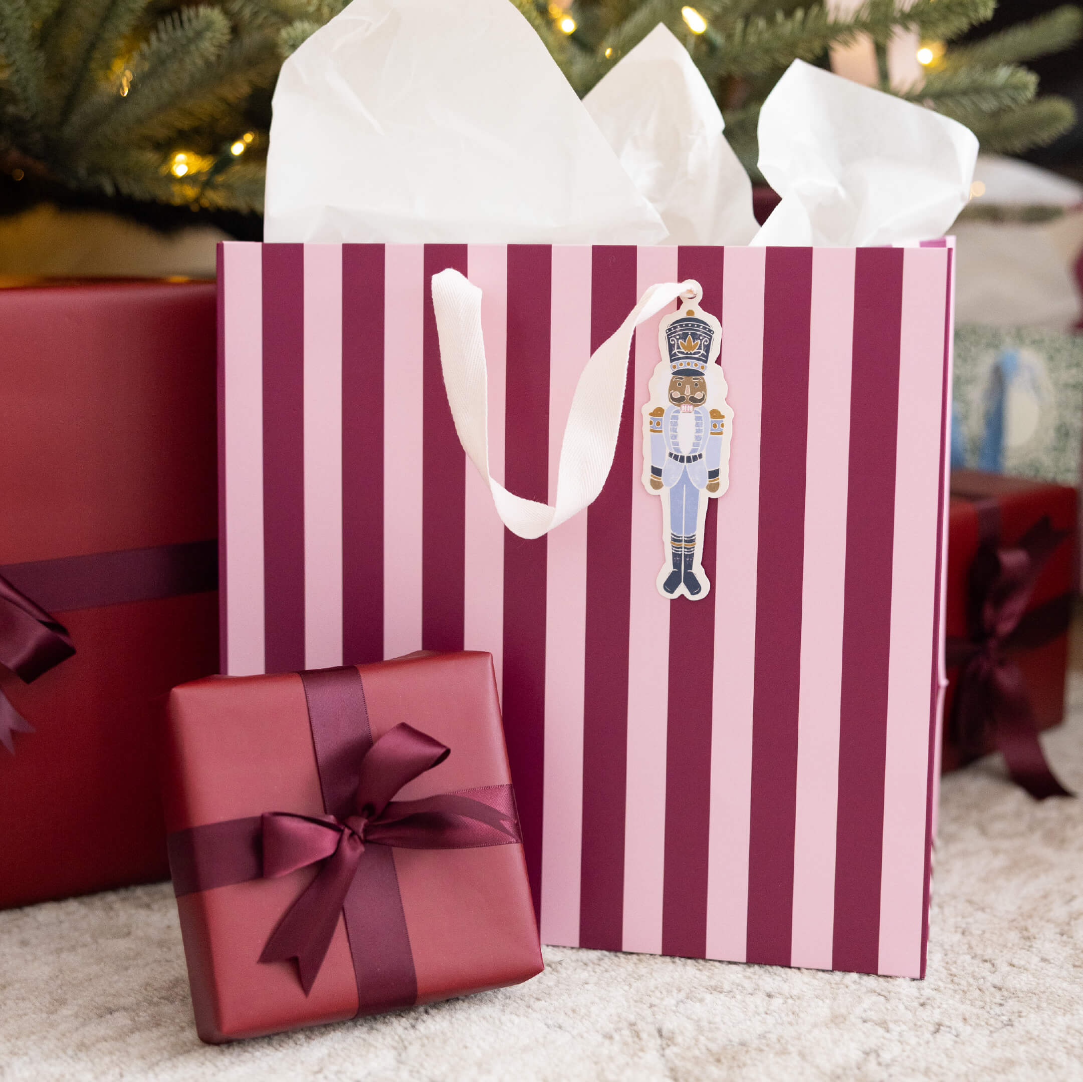 Berry Stripe Large Gift Bag with Nutcracker gift tag under a Christmas tree beside presents wrapped with burgundy ribbon