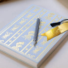A closed Simplified Linen Journal - Block Floral with a blue and gold floral cover, a black-and-white striped pen, and a gold tassel bookmark rest on a light surface.