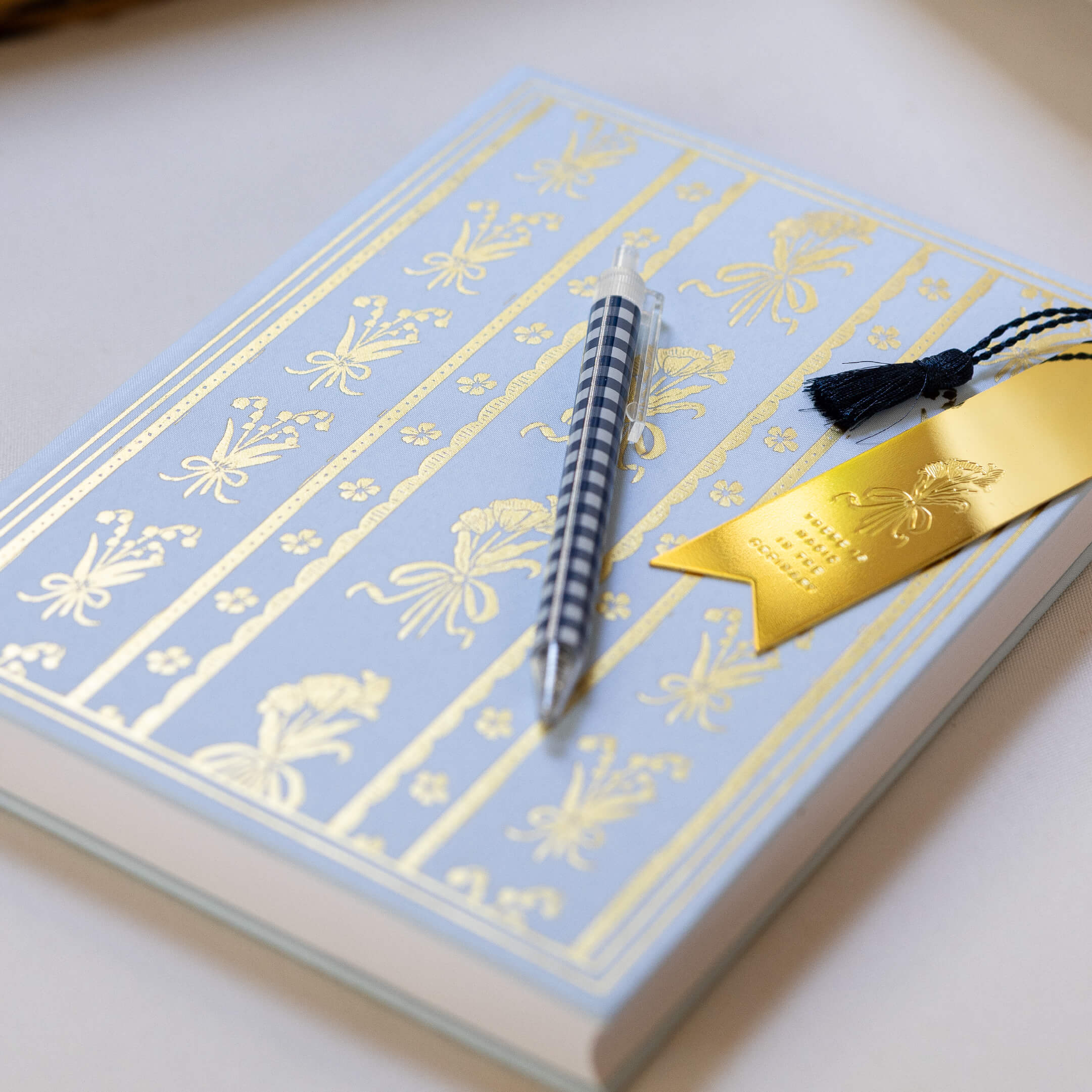 A closed Simplified Linen Journal - Block Floral with a blue and gold floral cover, a black-and-white striped pen, and a gold tassel bookmark rest on a light surface.