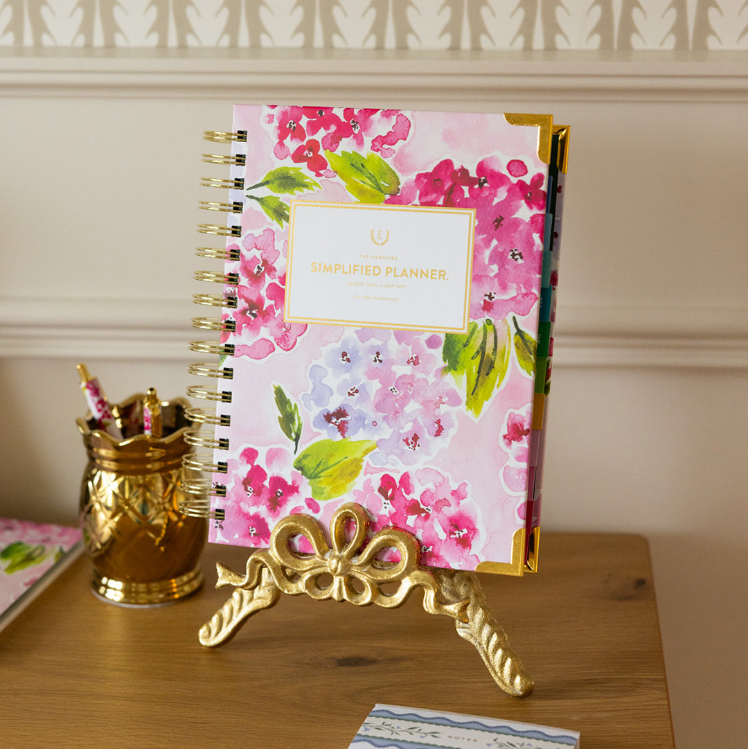 Simplified by Emily Ley floral planner displayed on desk with gold stand, pink watercolor cover, and elegant workspace styling