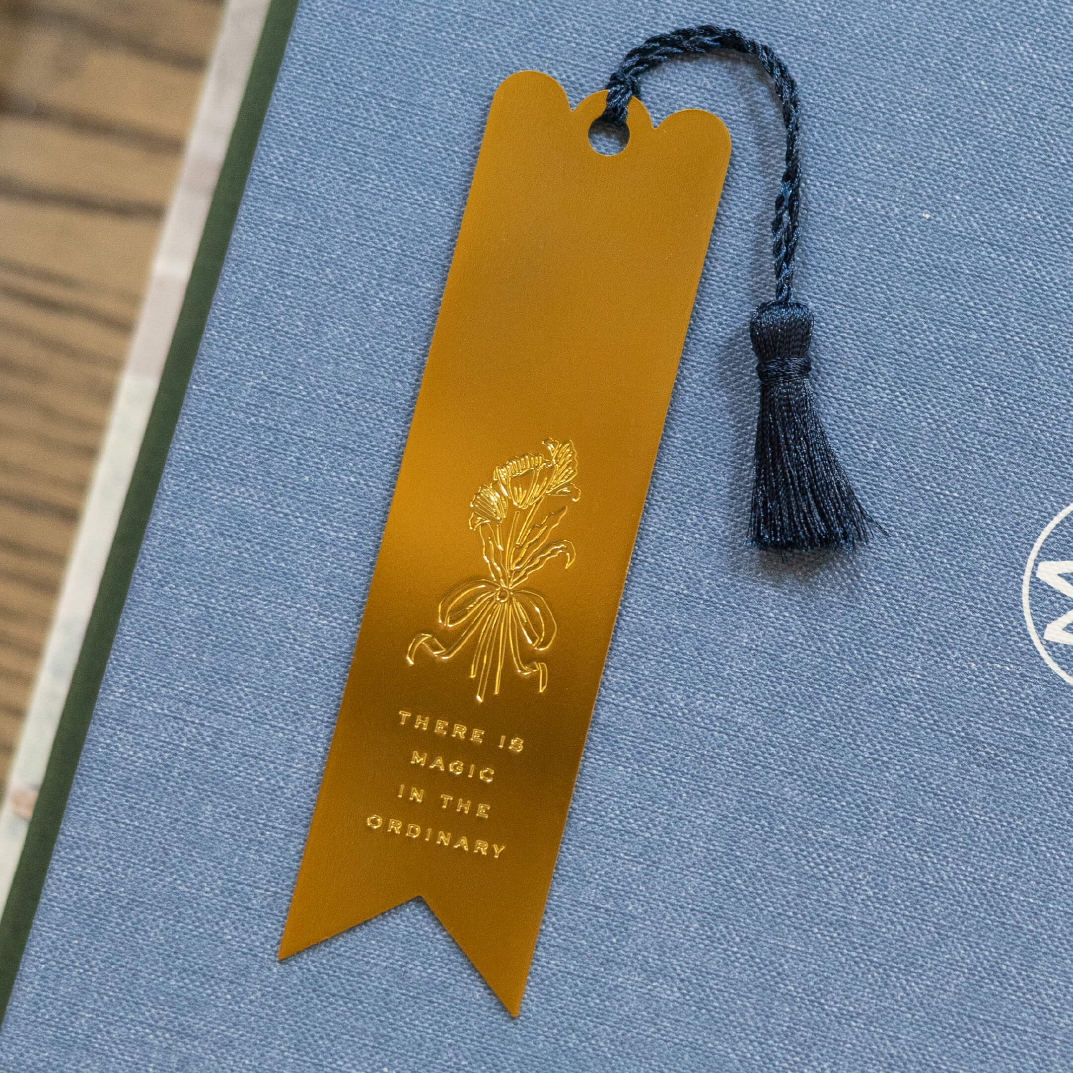 The Simplified Engraved Bookmark - Magic In The Ordinary features a delicate floral design and the phrase There is magic in the ordinary, with a gold finish, navy blue tassel, and rests beautifully on a grayish-blue book cover.