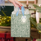 Emerald Floral Medium Gift Bag with green leaf print and light blue ribbon handles, held by a hand near a Christmas tree