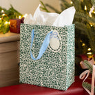 Emerald Floral Medium Gift Bag with white tissue paper, light blue ribbon handles, and a bow gift tag by a Christmas tree