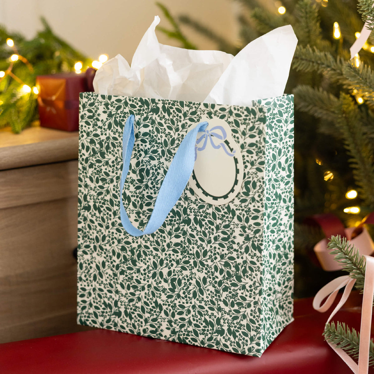Emerald Floral Medium Gift Bag with white tissue paper, light blue ribbon handles, and a bow gift tag by a Christmas tree