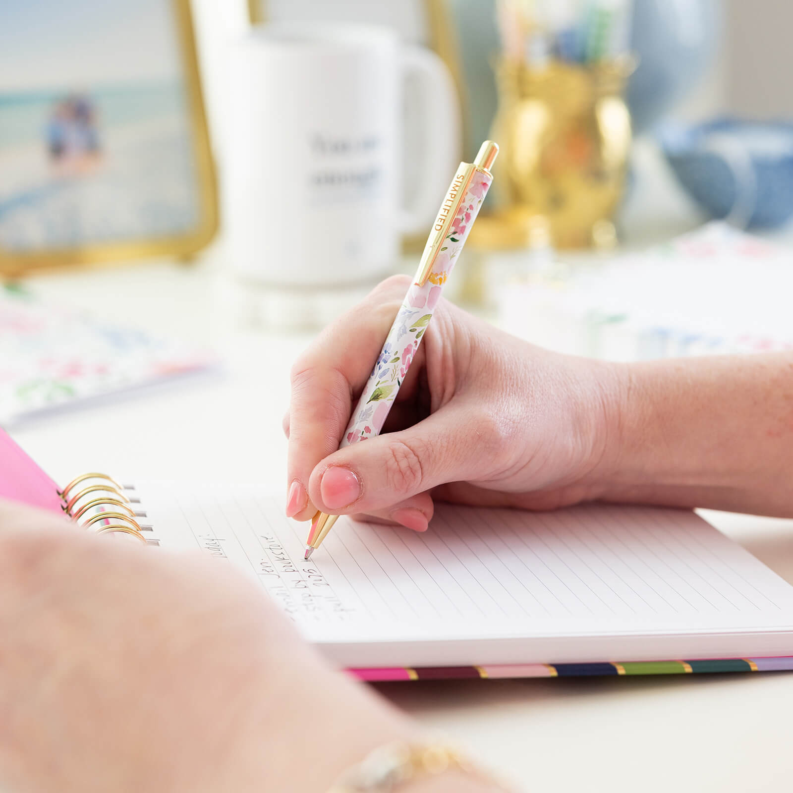 Someone writes in a Simplified Click Pen - Cottage Floral spiral notebook. On the desk: a mug, decorative container, and blurred photos—an ideal everyday writing essential for anyone.