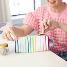 At a desk, someone in a pink textured top opens the Simplified Pen Case - Happy Stripe, featuring colorful vertical stripes and a leatherette exterior. A small jar of paper clips sits nearby.