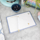 The Simplified Meal Planning Notepad with a pen rests on a marble surface, ready for planning meals and grocery lists, beside blueberries, books, and a potted plant.