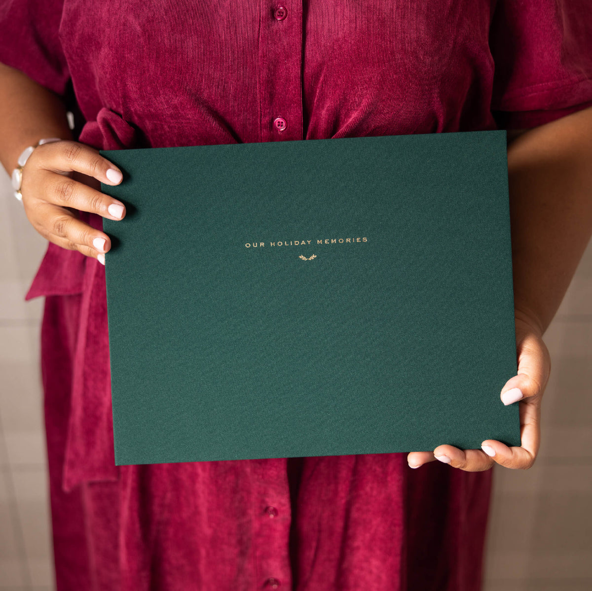Evergreen Holiday Memories Book in green linen with a gold foil Our Holiday Memories title, held against a burgundy dress
