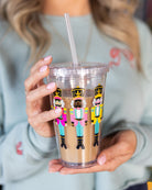 A person in a light blue top holds the Simplified Clear Tumbler - Nutcrackers, a 16oz insulated tumbler with a lid and straw, featuring colorful nutcracker designs and filled with a light brown iced drink.