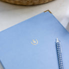 The Simplified Leatherette Folio in French Blue, featuring a gold emblem, sits on a white surface next to a blue and white checkered pen. A woven basket appears in the background, creating an elegant workspace scene.