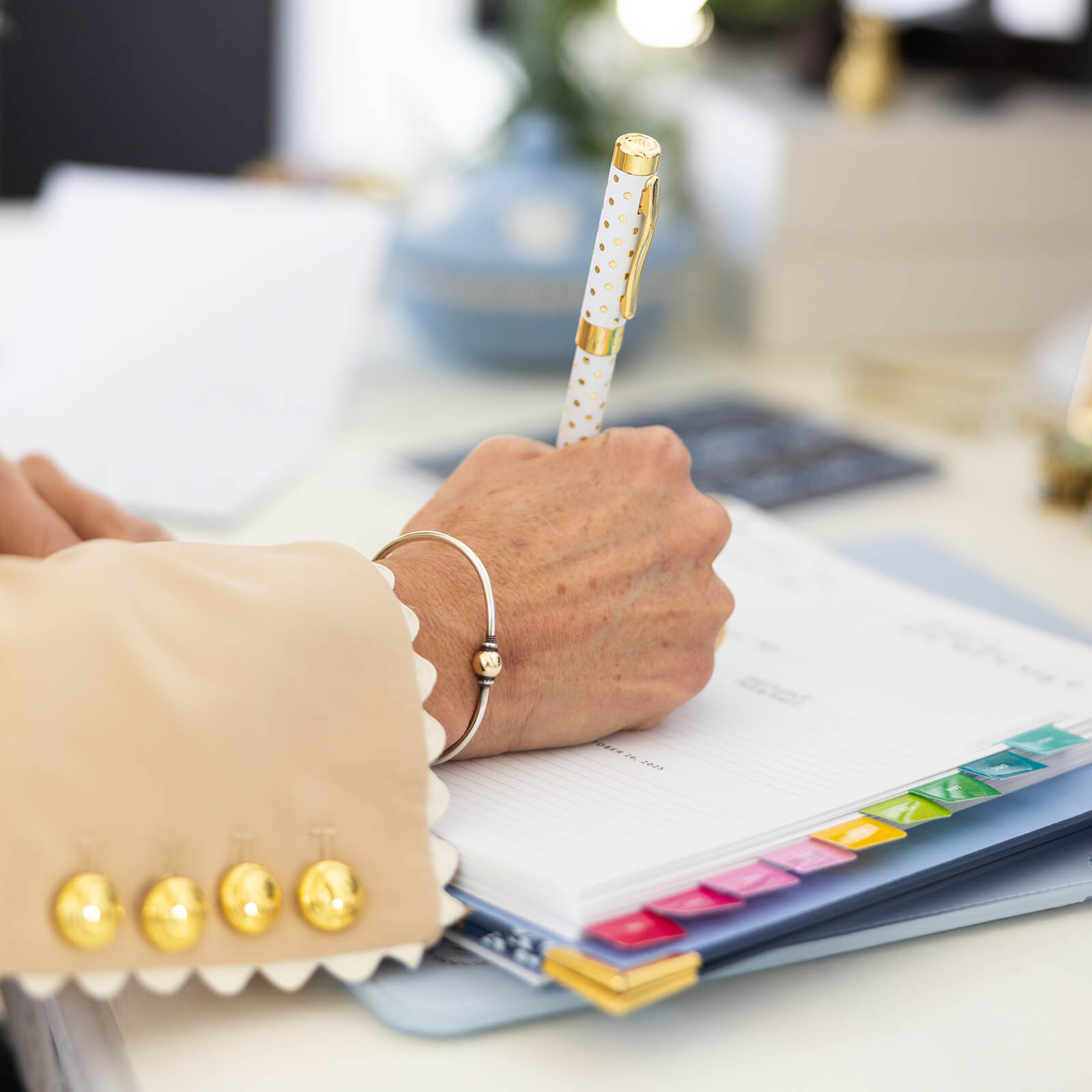 A person wearing a beige jacket with gold buttons writes in a planner using the Simplified Pen - Gold Dot. The planner features colorful tab dividers and rests on a desk, with office items blurred in the background.