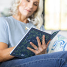 A blonde woman, dressed in a light blue shirt and jeans, reflects and writes creatively in the Simplified Linen Journal - Navy, enjoying self-expression with this elegant journal in her hands.