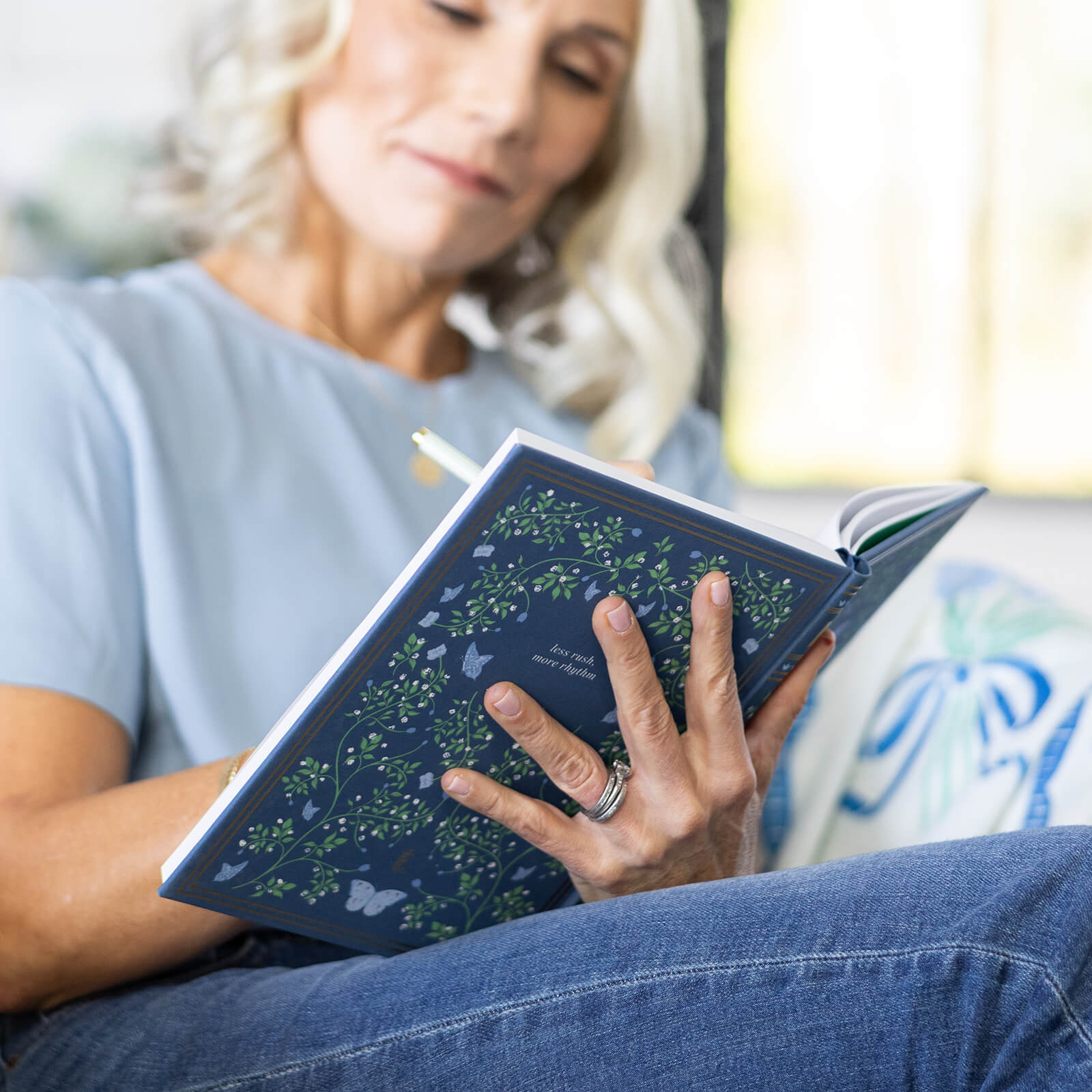 A blonde woman, dressed in a light blue shirt and jeans, reflects and writes creatively in the Simplified Linen Journal - Navy, enjoying self-expression with this elegant journal in her hands.