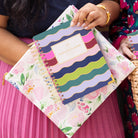 Two people hold the Simplified Planner Pouch - Cottage Floral with gold zipper teeth and a vibrant spiral-bound planner. One wears a pink pleated skirt; the other is dressed in floral patterns.