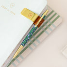 A 2024-2025 planner with a gold clip holds a floral pen with gold accents, resting on a striped desk beside Simplifieds Misty Meadow mechanical pencil featuring a sleek metal body.