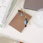 A pair of glasses rests atop the Simplified Sticker Book - Dapperdesk, packed with 2,000+ stickers, on a white desk near a planner, closed laptop, and glass of ice water.