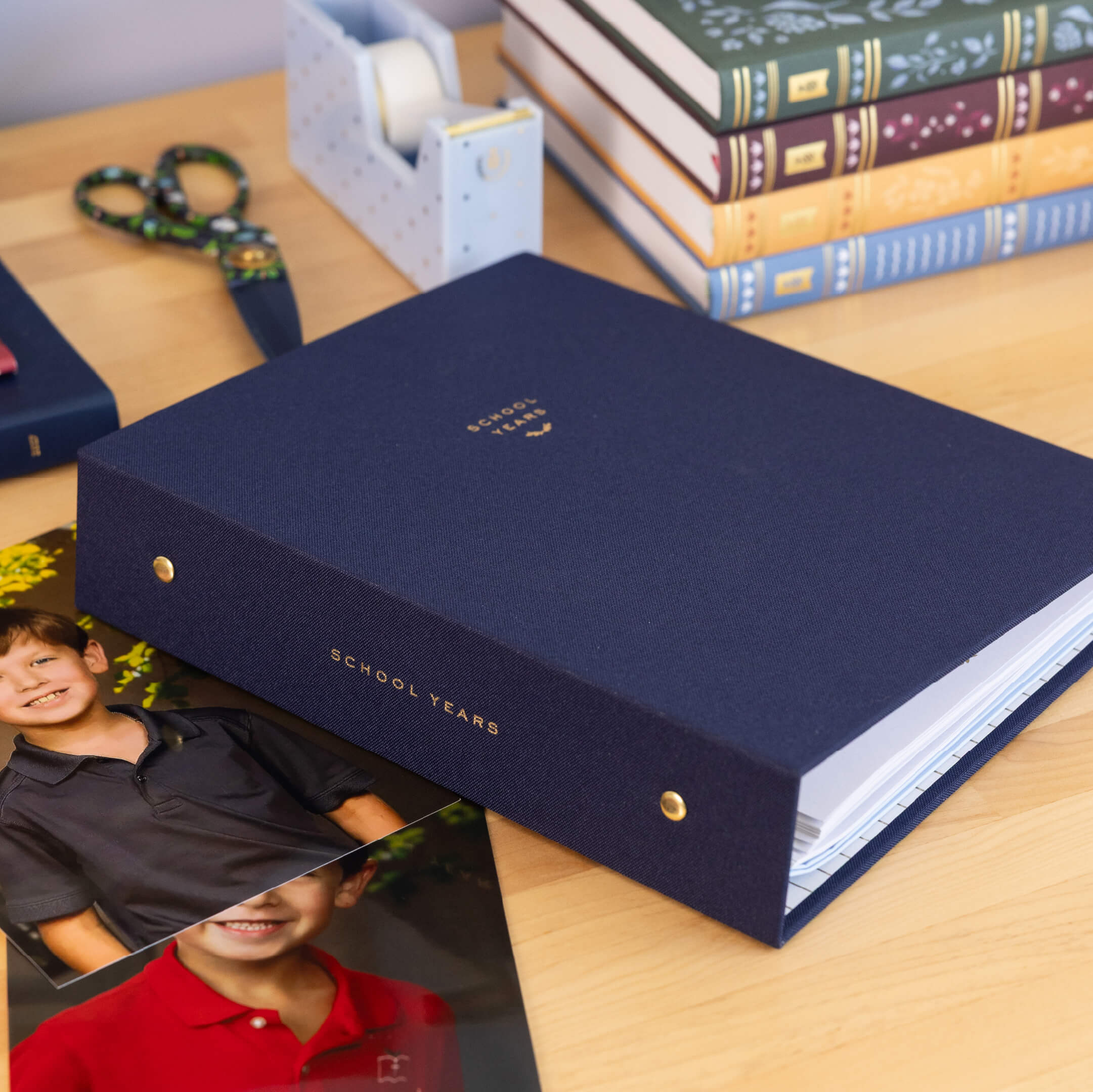 Navy linen School Years Book on a wooden desk with school portrait photos, floral scissors, a tape dispenser and a stack of journals, showing the gold foil spine and brass snap details.