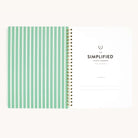 A spiral-bound notebook with left pages featuring green lines and a right page titled Workbook - Finances by Simplified. This money management tool includes a Belongs to section at the bottom to help you achieve your financial goals.