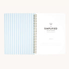 An open spiral-bound wellness workbook by Simplified is shown. The left page displays blue and white stripes, while the right page invites goal tracking with a Belongs To section beneath the title Workbook - Wellness.